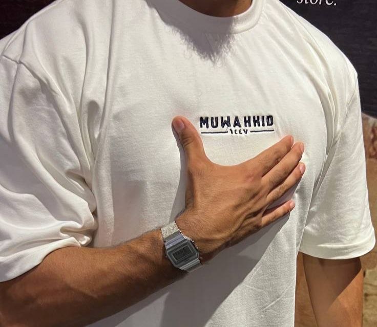 A close-up of a person wearing a white oversized T-shirt with the text 'MUWAHID UCEY' printed in black across the chest, with one hand resting on the shirt. A silver digital watch is also visible on the wrist.